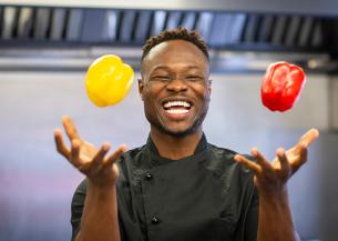 A Sanctuary Care chef wearing chef blacks juggling with a yellow and red pepper whilst laughing