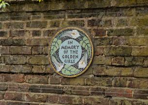 Circular mosaic plaque on a brick wall with the text 'IN MEMORY OF THE GOLDEN GIRLS', surrounded by white birds and green leaves