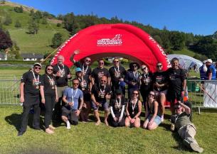 A group of people wearing black 'Walking With The Wounded' t-shirts pose in front of a red inflatable arch with the same logo. They are outdoors on grass with hills and trees in the background. Some are standing, kneeling, or sitting, and many are holding medals.