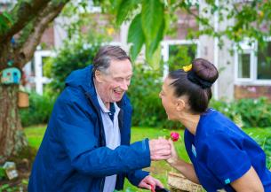 Person in blue jacket holds pink flower while another in blue scrubs leans forward; both are outdoors near a tree and building with greenery.