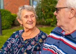 Two elderly individuals are sitting outdoors on a bench, facing each other. One wears a dark floral blouse with a gold necklace, and the other wears a blue polo shirt with red, white, and blue stripes. They appear to be engaged in conversation. The background includes greenery and part of a brick building. 