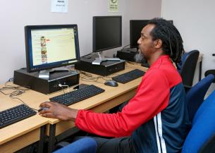 A man with long, curly hair and a beard is sitting at a computer desk in a room with multiple computers. He is wearing a red and black athletic jacket and is focused on the screen in front of him, which displays a web browser. The workspace is simple, with wooden desks and blue chairs.