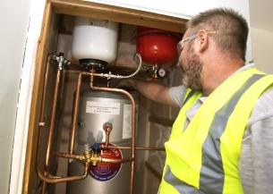 A technician wearing a safety vest and glasses is inspecting a water heater installation. The water heater is cylindrical and metallic, with various copper pipes connected to it. The technician is pointing at a component on the heater, which is mounted in a wooden enclosure. The background shows unfinished walls, indicating that the area is still under construction.