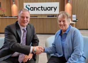 Two people are seated in modern chairs in a reception area, shaking hands. Behind them is a wooden-panelled wall with a large sign that reads “Sanctuary.”