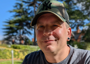 Sanctuary customer Alan Tyla looks towards the camera and smiles, He is sat outdoors in a grassy area and wearing a t-shirt and baseball cap.