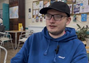 Person in blue Levi’s hoodie and black cap sitting in a café with bulletin board and chairs in background.