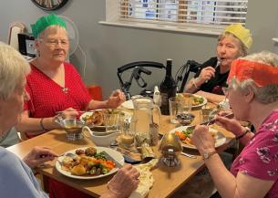 A group of four elderly women sitting around a table enjoying a meal together. They are wearing colorful paper crowns and are engaged in conversation.