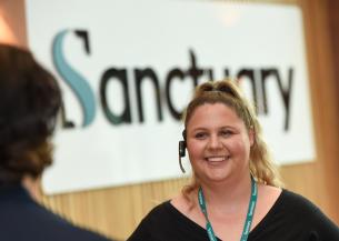 A smiling woman with curly blonde hair is wearing a headset and standing in front of a sign that reads "Sanctuary."