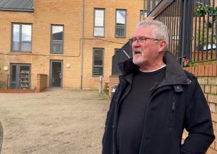 A man with grey hair and beard stands in front of a modern brick block of flats
