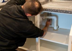 A person wearing a black hoodie and work pants is crouched down under a kitchen sink, working on the plumbing. The sink has a stainless steel surface and a faucet, and there are cleaning supplies and paint cans visible on the countertop.