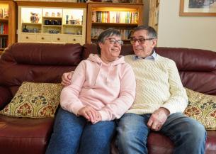A middle-aged couple sits closely together on a comfortable sofa, smiling at each other. The woman wears a light pink hoodie and glasses, while the man is dressed in a cream-colored sweater and glasses. They are surrounded by a cozy living room with bookshelves and decorative items in the background. The atmosphere is warm and affectionate.