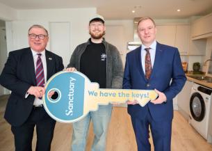 Three men are standing in a modern kitchen. The man in the middle is holding a large, symbolic key with the word "Sanctuary" and the phrase "We have the keys!" printed on it.
