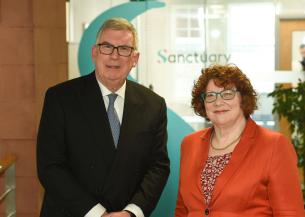 Two people standing side by side in an office reception area, with a large Sanctuary logo visible behind them.