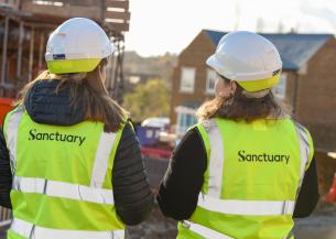 Two women wearing safety helmets and high-visibility vests with the word "Sanctuary" printed on the back are standing with their backs to the camera. They are on a construction site, with scaffolding and partially constructed buildings visible in the background. The scene is illuminated by sunlight, suggesting a clear day.