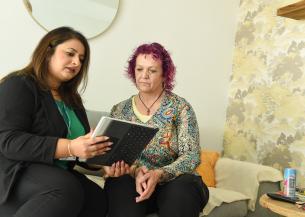 Colleague sits with a customer in a living room, using a tablet to review information together. The customer is seated on a sofa while the colleague points to items on the screen during a home visit.