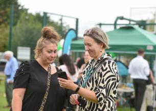 Two women are standing outdoors, engaged in conversation while looking at a smartphone. One woman, with a bun hairstyle and wearing a black top, appears to be listening intently. The other woman, with short grey hair and dressed in a patterned black and white outfit, is showing something on the phone. In the background, there are people mingling and green tents set up, indicating a social event or gathering.
