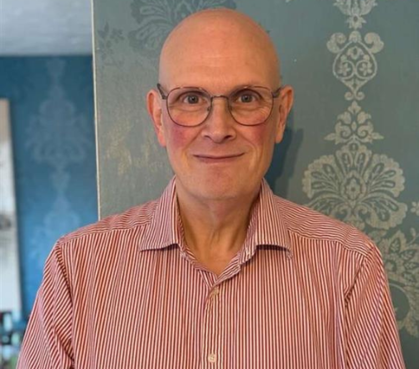 A smiling, bald man wearing round glasses stands against a wall decorated with pale blue wallpaper