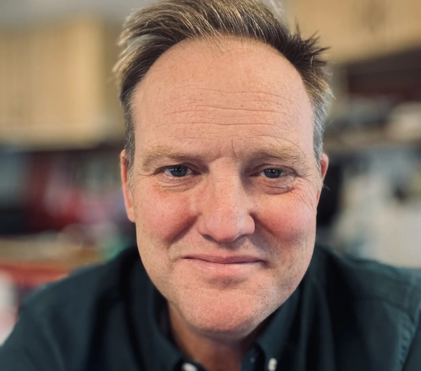 A head and shoulders photo of Tristram Channer today, working for Sanctuary. He is looking towards the camera and smiling. He has short, light brown hair and is wearing a dark button-up shirt.