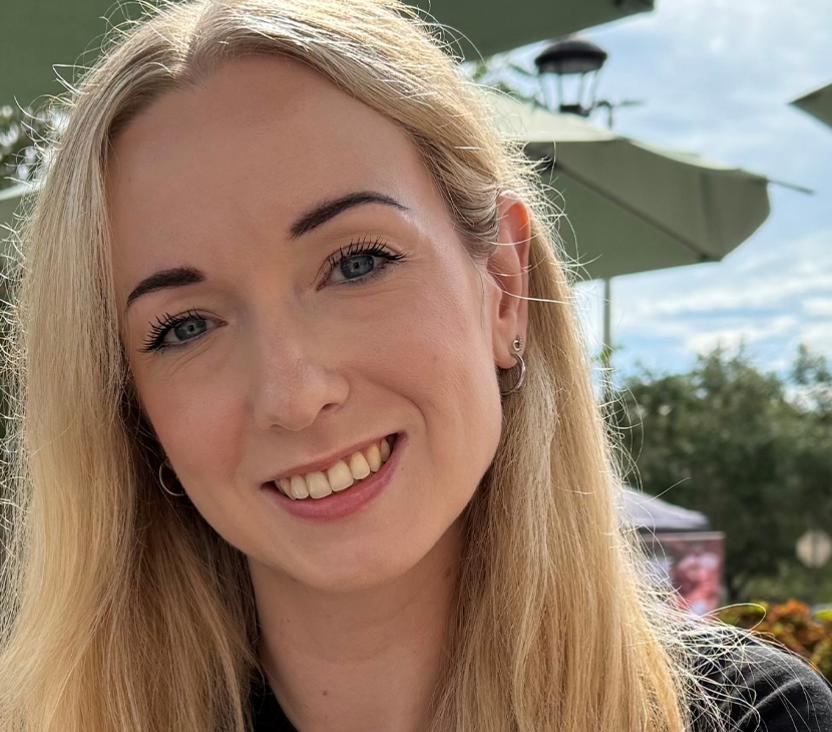 A head and shoulders photo of a woman with long blonde hair. She is wearing a smart black t-shirt and smiling towards the camera.