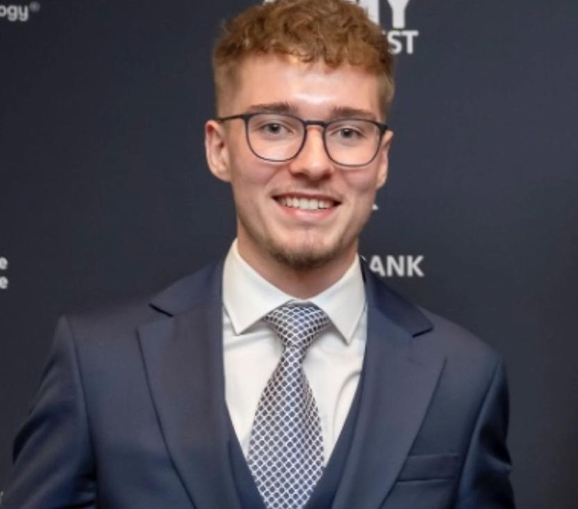 A man in a navy blue suit and patterned tie looks towards the camera and smiles. He has short brown hair and is wearing glasses.